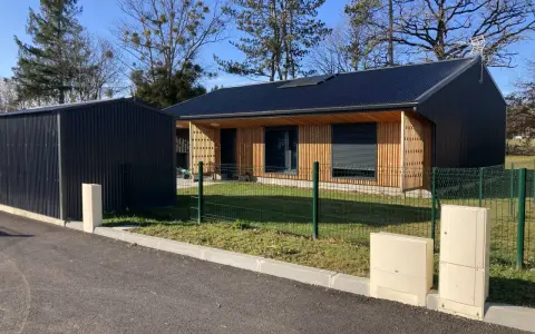 Maison passive ossature bois avec panneaux solaires sur le toit, bardage bois et métal, devant un ciel bleu et des arbres.