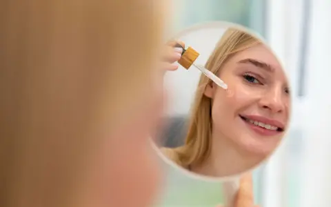 Mujer rubia sonríe mientras se aplica aceite facial con gotero en la mejilla. Un paso esencial en su rutina de cuidado de la piel, ya sea aceite facial antes o después de la crema.
