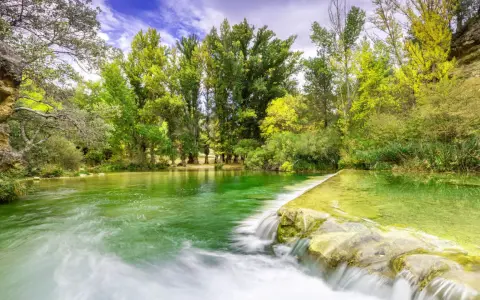 La poza de doña urraca, un remanso de agua verde esmeralda con una pequeña cascada, rodeada de frondosa vegetación y árboles altos bajo un cielo azul.