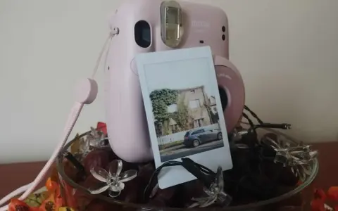 Różowy aparat Instax z wydrukowanym zdjęciem domu i samochodu. Idealny pomysł, jak zrobić prezent ze zdjęć, ozdobiony jesiennymi liśćmi i lampkami.