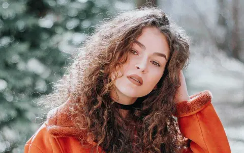 Jeune femme aux cheveux bouclés, portant une veste orange. La retouche photo netteté en ligne sublime son regard intense.