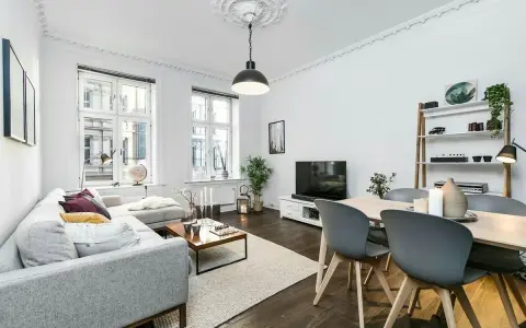 Elegante salón con sofá gris, mesa de centro y comedor. El **suelo oscuro** de madera contrasta con las paredes blancas y la luz natural.