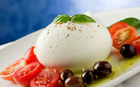 Duża kulka mozzarelli di bufala z pomidorkami koktajlowymi, oliwkami i bazylią na białym talerzu.
