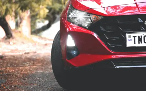 Czerwony Hyundai i20 z zapalonymi światłami dziennymi i reflektorami. Widać fragment maski, grill i tablicę rejestracyjną.