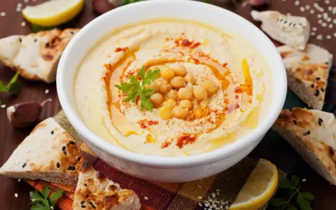 Un bol de houmous maison, garni de pois chiches, de persil et de paprika, servi avec du pain pita, du citron et de l'ail.