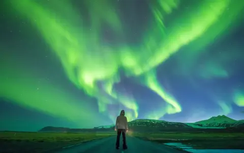 Człowiek stoi na drodze pod rozgwieżdżonym niebem, gdzie tańczy zorza polarna. Islandia, październik, pogoda idealna na takie widowisko.