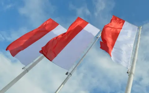 Trzy powiewające na wietrze flagi, każda z czerwono-białą szarfą, na tle błękitnego nieba z chmurami.