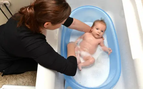 Mama delikatnie myje noworodka w niebieskiej wanience. Maluch pluska się w pianie, a mama uśmiecha się do niego.