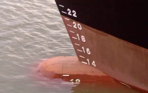 La coque d'un navire, marquée de -22 à -14, flotte sur l'eau. Le déplacement léger du bateau est visible.