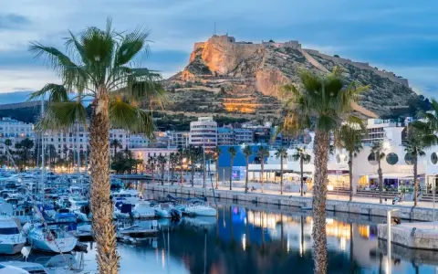 Palme e barche nel porto di Alicante, con il Castello di Santa Barbara illuminato sullo sfondo.
