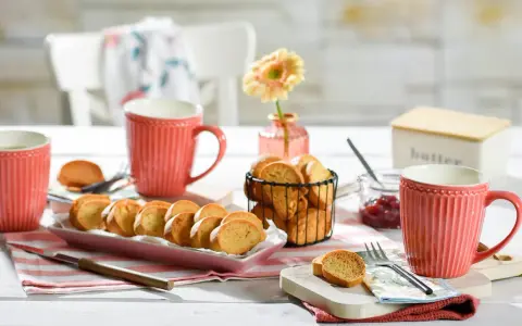 Leckeres Zwieback-Rezept: goldbraune Scheiben auf rosa Platte, dazu Marmelade, Butterdose und rote Tassen.