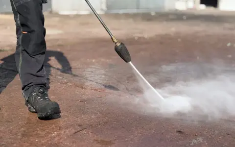 Osoba w roboczych spodniach i butach czyści mokrą nawierzchnię za pomocą myjki ciśnieniowej z dyszą rotacyjną.