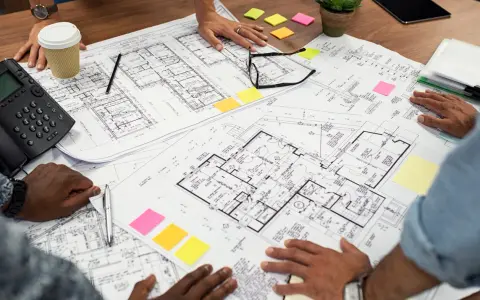 Des mains examinent un plan d'une maison, avec des post-it colorés et un téléphone sur la table.