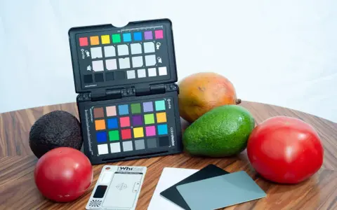 Na drewnianym blacie leżą owoce: awokado, mango, pomidory. Obok znajduje się otwarty colorchecker z kolorowymi polami i szara karta do kalibracji balansu bieli.