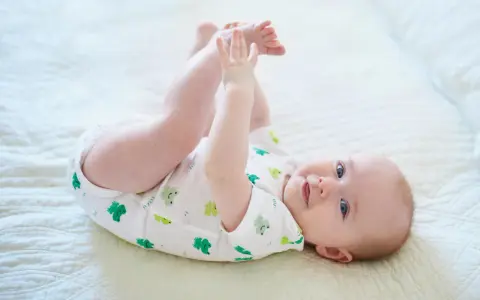 Ein süßes Baby, ca. 6 Monate alt, liegt auf dem Rücken und spielt mit seinen Füßen. Es trägt einen weißen Body mit grünen Fröschen.