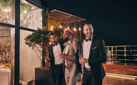 Trzech eleganckich panów, w tym drużba na weselu, śmieje się i trzyma drinki na tarasie nocą, wśród ozdobnych świateł.