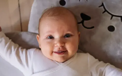 Uśmiechnięte niemowlę w białym body leży na miękkiej poduszce w kształcie misia. To mądre imię dla chłopca, które przyniesie mu szczęście.