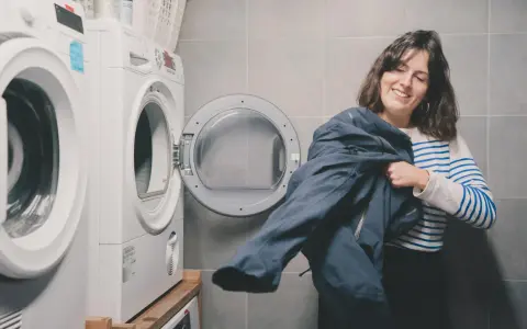 Kobieta z uśmiechem trzyma granatową kurtkę, przygotowując się do prania Gore-Tex. Obok stoi otwarta suszarka.