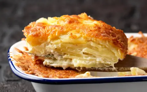 Une part généreuse de gratin de pomme de terre, avec sa croûte dorée et ses couches crémeuses, est soulevée par une spatule en bois.