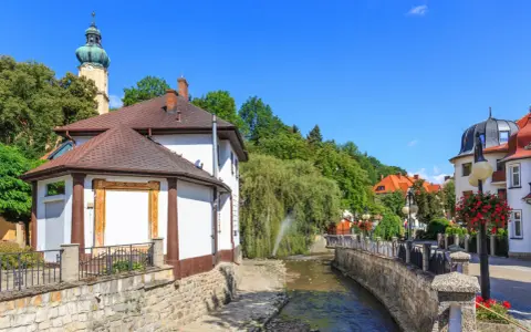 Urokliwy strumyk w Polanicy Zdrój, otoczony kamiennymi murkami i zielenią. W tle widać wieżę kościelną i kolorowe kamieniczki.