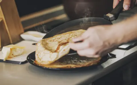 Une main retourne une crêpe dorée dans une poêle fumante. Sur le côté, du beurre frais attend. Ces meilleures crêpes promettent un délice.