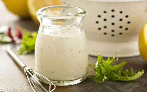 Ein Glas cremiges Ranch Dressing, daneben ein Schneebesen, Salatblätter und Zitronenspalten. Perfekt für dein Ranch Dressing Rezept!
