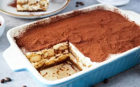 Ein köstliches Tiramisu rezept ohne alkohol, bestäubt mit Kakao, in einer blauen Auflaufform. Ein Stück ist bereits entnommen.