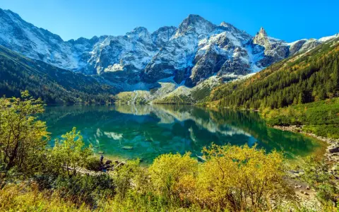 Widok na Morskie Oko z wysokości. Turkusowa tafla jeziora odbija ośnieżone szczyty Tatr. Jesienne drzewa otaczają brzeg.