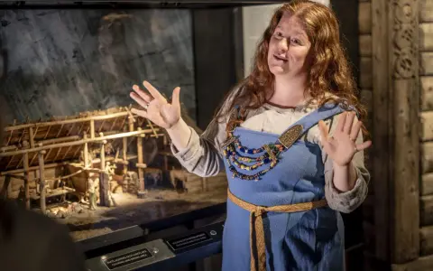 En kvinna i vikingakläder berättar om en modell av en vikingagård på ett vikingamuseum.