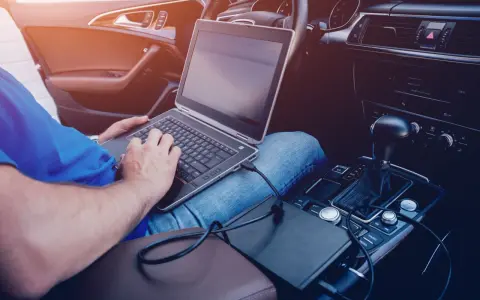 Mężczyzna w samochodzie podłącza laptopa do zasilania, pokazując, jak podłączyć komputer do samochodu.
