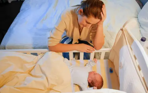 Zmęczona mama pochyla się nad łóżeczkiem, zastanawiając się, kiedy nastąpi regres snu. Niemowlę śpi spokojnie.