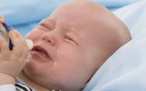 Ein weinendes Baby mit geschlossenen Augen, dem eine Flasche angeboten wird. Das Baby will nicht trinken.