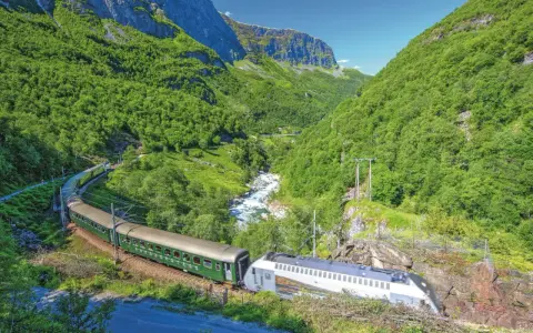 Grüner Zug fährt durch ein bergiges Tal mit Fluss. Die Fahrt von Bergen nach Oslo ist ein unvergessliches Erlebnis.