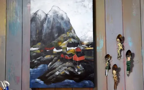 Målade landskap med röda hus vid en bergig kust. Konstverket hänger på en vägg av slitna träplankor.