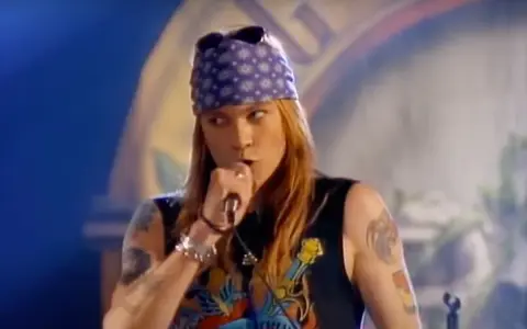 Axl Rose w fioletowej bandanie i czarnej koszulce śpiewa do mikrofonu, jakby dedykował "sweet child o' mine" publiczności.