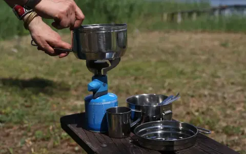 Na stole piknikowym stoi menażka, naczynie turystyczne, obok kubek i talerz.