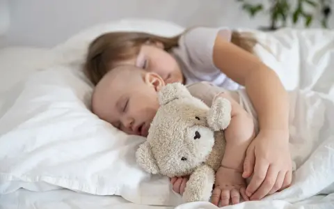 Dzieci śpią wtulone w misia. Starsza siostra czuwa nad maluszkiem, pokazując, ile śpi dwulatek i jak ważny jest sen dla rozwoju.