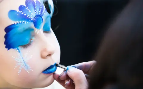 Ein Kind wird mit blauem Lidschatten und Schneeflocken bemalt, um wie Elsa auszusehen. Das Schminken ist leicht und zaubert ein magisches Wintergesicht.