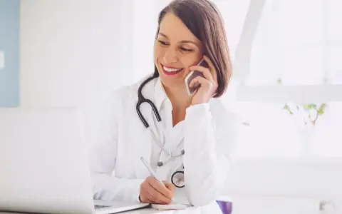 Lekarka w białym kitlu rozmawia przez telefon i pisze notatki przy laptopie.