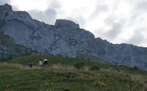 Randonneurs sur une pente herbeuse face à une falaise imposante. Le chemin sinueux et la hauteur suggèrent que la tournette est dangereuse.