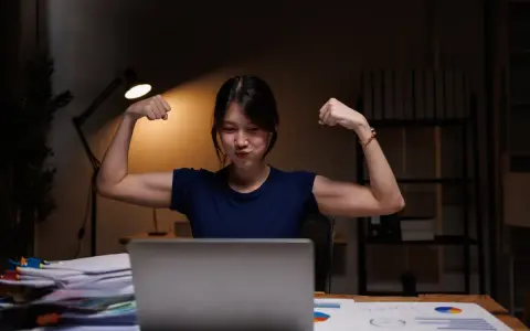Młoda kobieta w godzinach nocnych 2026, siedząc przy biurku, pokazuje bicepsy.