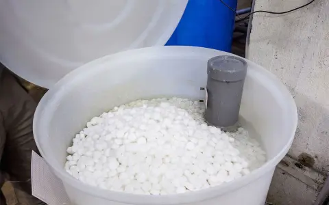 Duży biały pojemnik wypełniony tabletkami soli do zmiękczacza wody. W środku widoczny szary element instalacji.