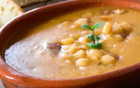 Una ciotola di terracotta piena di zuppa di fagioli cremosa, con pezzetti di carne e una fogliolina di menta. Perfetta per una cena confortante.