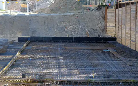 Armatura per fondazione a platea sezione. La rete metallica è pronta per il getto di calcestruzzo, con terra e muri di contenimento sullo sfondo.