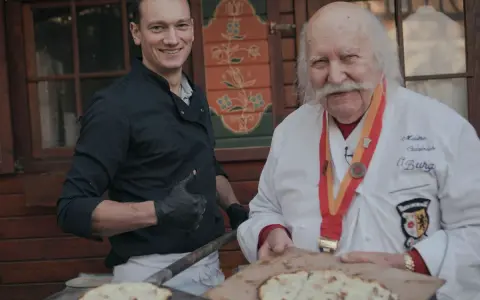 Deux chefs, l'un jeune et souriant, l'autre âgé avec une médaille, présentent des tartes flambées, évoquant l'origine du flamenkuch.