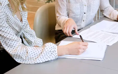 Two women in polka-dot shirts review documents. One takes notes, implying they are discussing who can see committee meeting minutes.