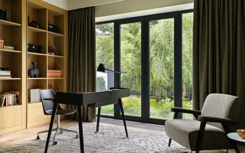 Acogedora habitacion despacho en casa con estanterías de madera, escritorio negro y sillón estampado. Ventanal con vistas al jardín.