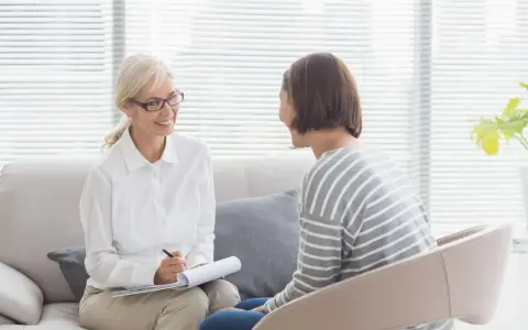 Psycholog a psychoterapeuta w przyjaznej atmosferze gabinetu. Kobieta w okularach notuje, druga słucha uważnie.
