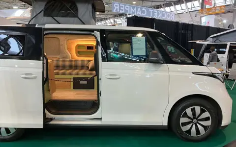 Vit ID Buzz camper van med uppfällt tak och öppen skjutdörr som visar en mysig interiör med rutig soffa och trädetaljer.