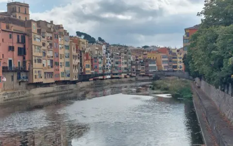 Bunte Häuserfronten säumen den Fluss Onyar in der malerischen **Girona Altstadt**. Eine Brücke verbindet die Ufer, während sich die Wolken am Himmel spiegeln.